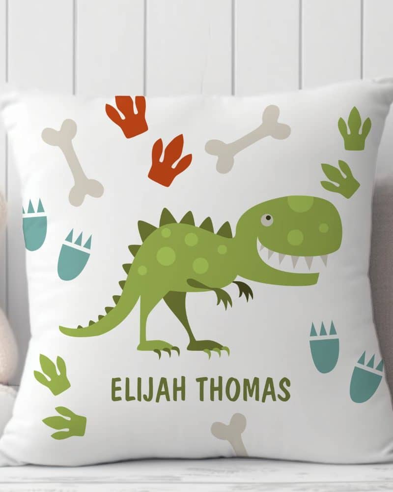 White pillow with a green dinosaur, dinosaur footprints, and bones. The name Elijah Thomas is printed below the dinosaur.