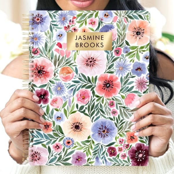Person Holding A Spiral-Bound Notebook With A Watercolor Floral Design And A Gold Nameplate.