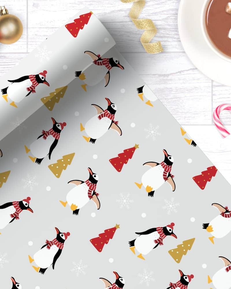 Grey wrapping paper with a repeating pattern of penguins wearing red scarves and hats, alongside small Christmas trees.