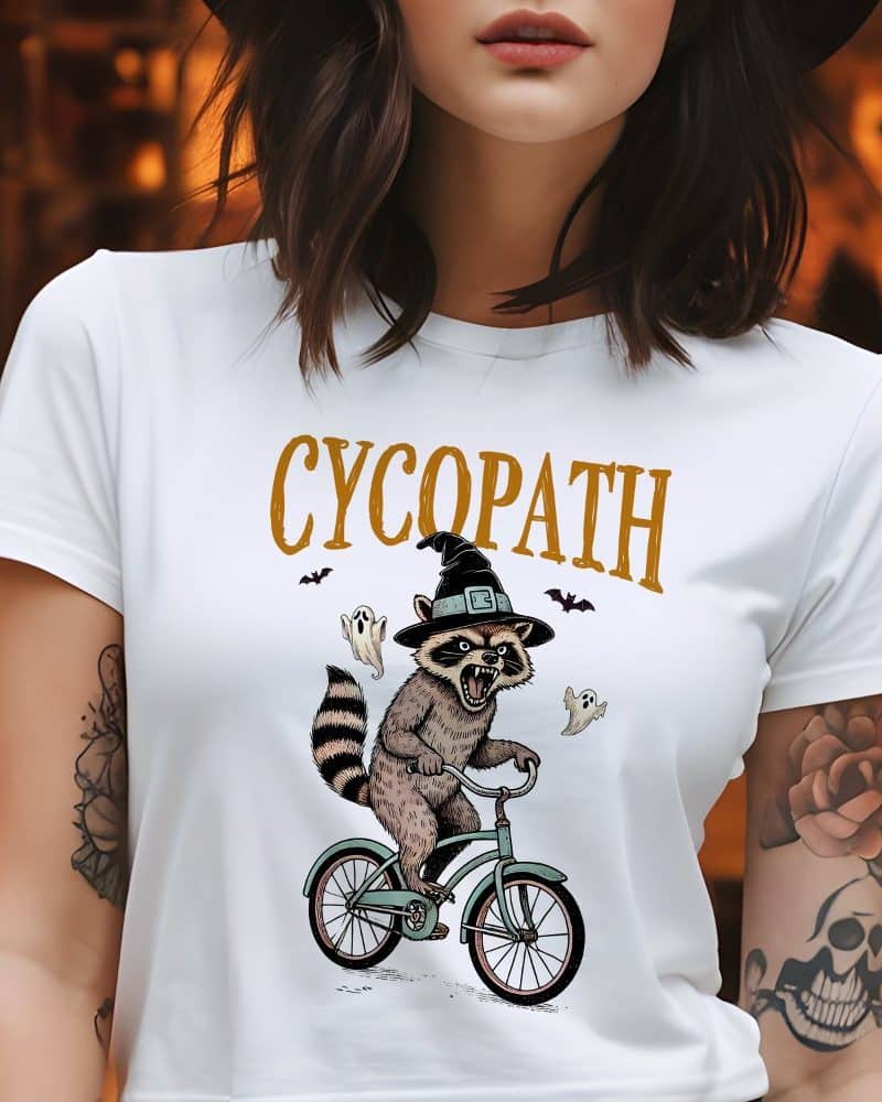White t-shirt featuring a cartoon raccoon wearing a witch's hat and riding a bicycle, surrounded by ghosts and bats; the word CYCOPATH is printed above.