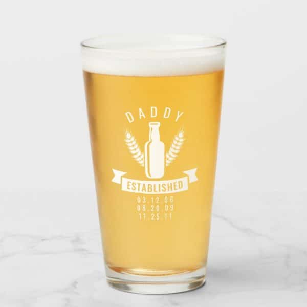 A Pint Glass Of Beer With A Design That Says Daddy Established And Lists Three Dates.