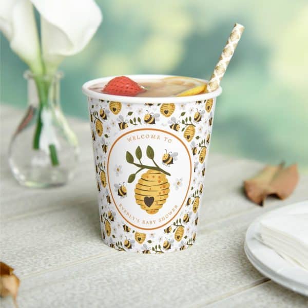 A Paper Cup With A Bee-Themed Baby Shower Design, Filled With A Strawberry Beverage, And A Gold Straw.