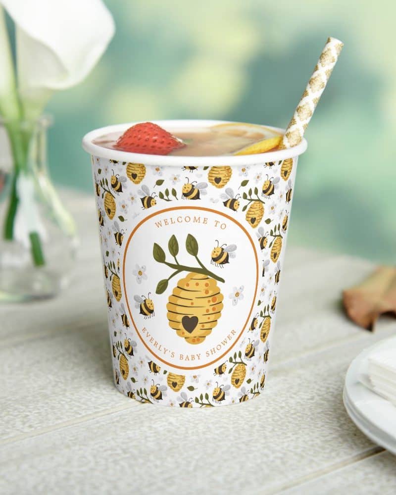A paper cup with a bee-themed baby shower design, filled with a strawberry beverage, and a gold straw.