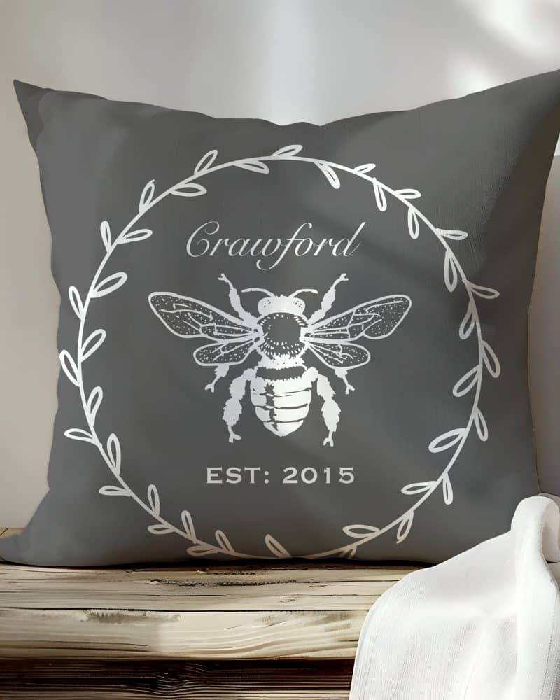 Gray pillow with a white bee illustration in a wreath, the name Crawford and the year 2015.