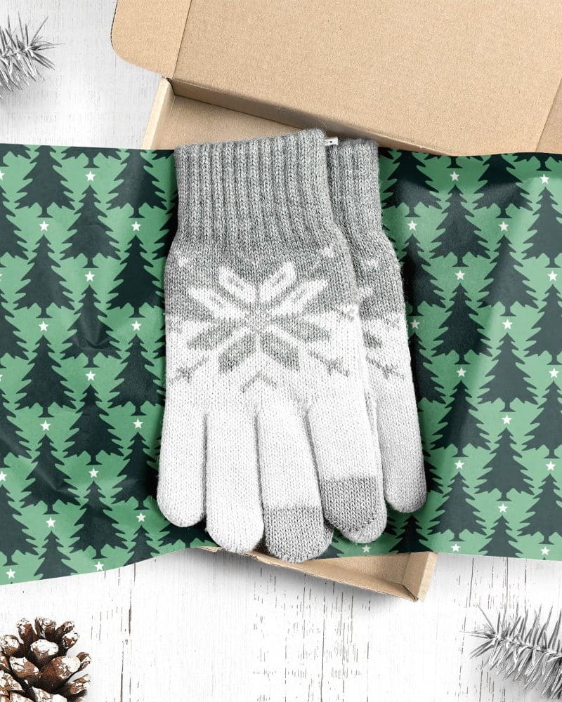 Pair of grey and white knit gloves with snowflake pattern, inside a box lined with Christmas tree patterned wrapping paper.