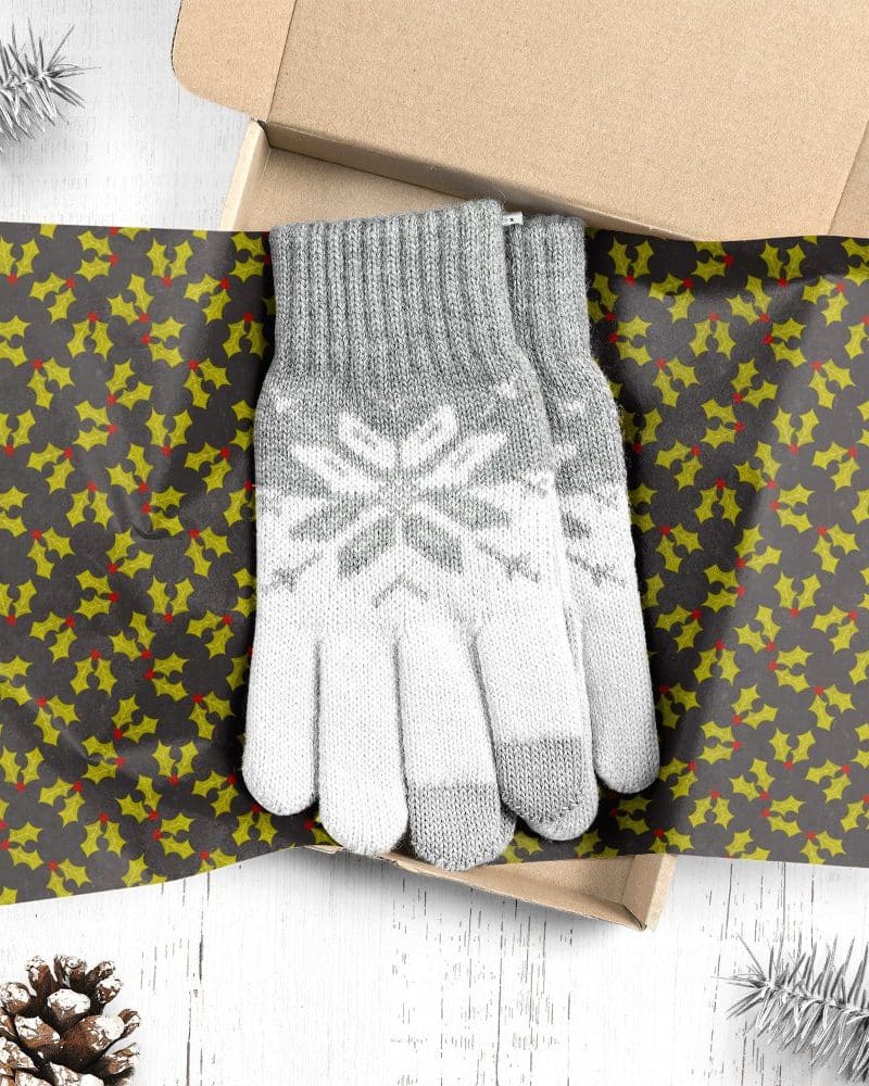 A pair of grey and white knit gloves with a snowflake pattern, in a gift box lined with holly-patterned wrapping paper.