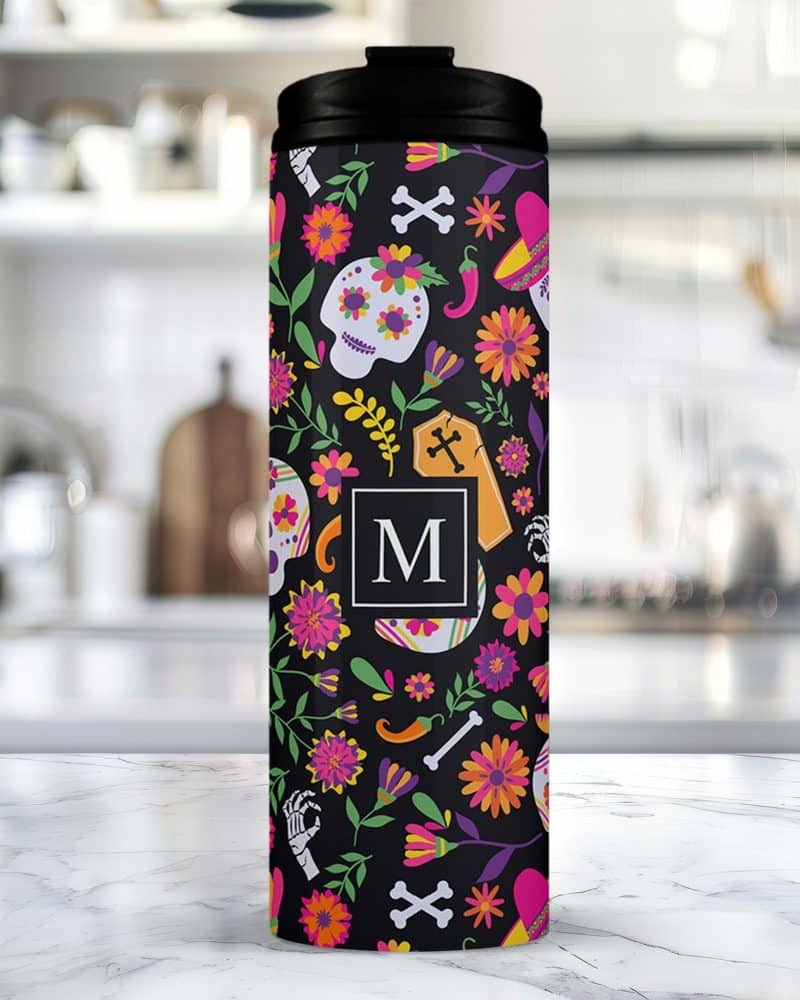 A black travel tumbler with a Day of the Dead design featuring sugar skulls, flowers, and a personalized initial.