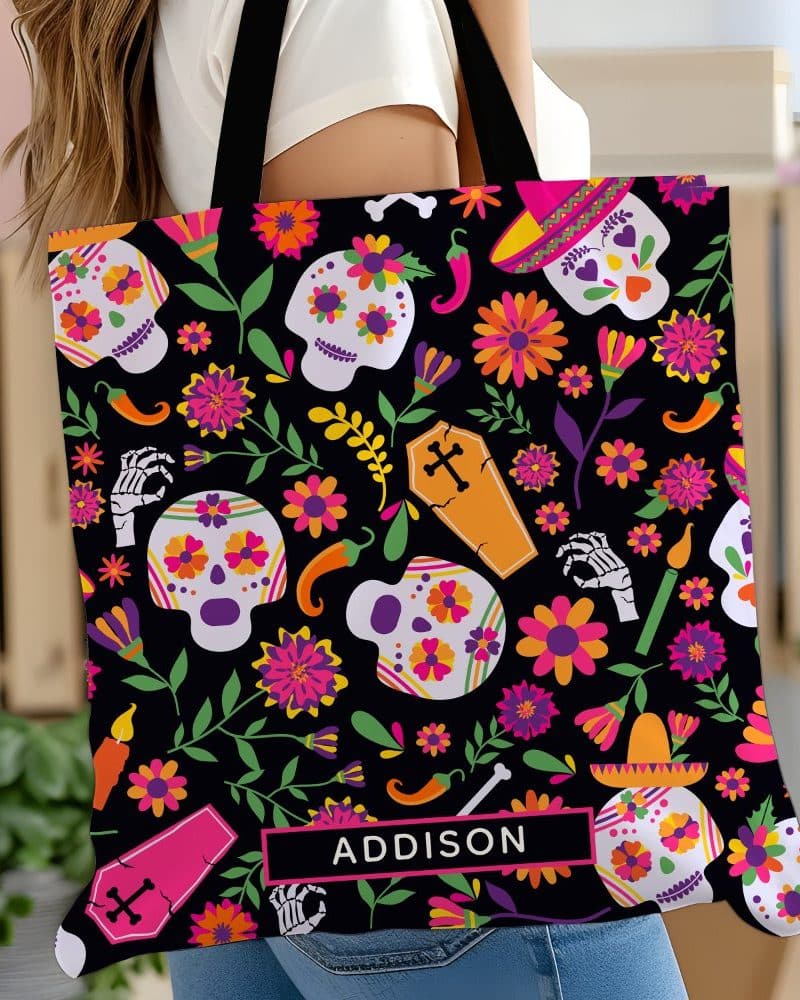 A tote bag with a Day of the Dead design featuring colorful sugar skulls, flowers, and other festive elements. The name Addison is printed on the bag.