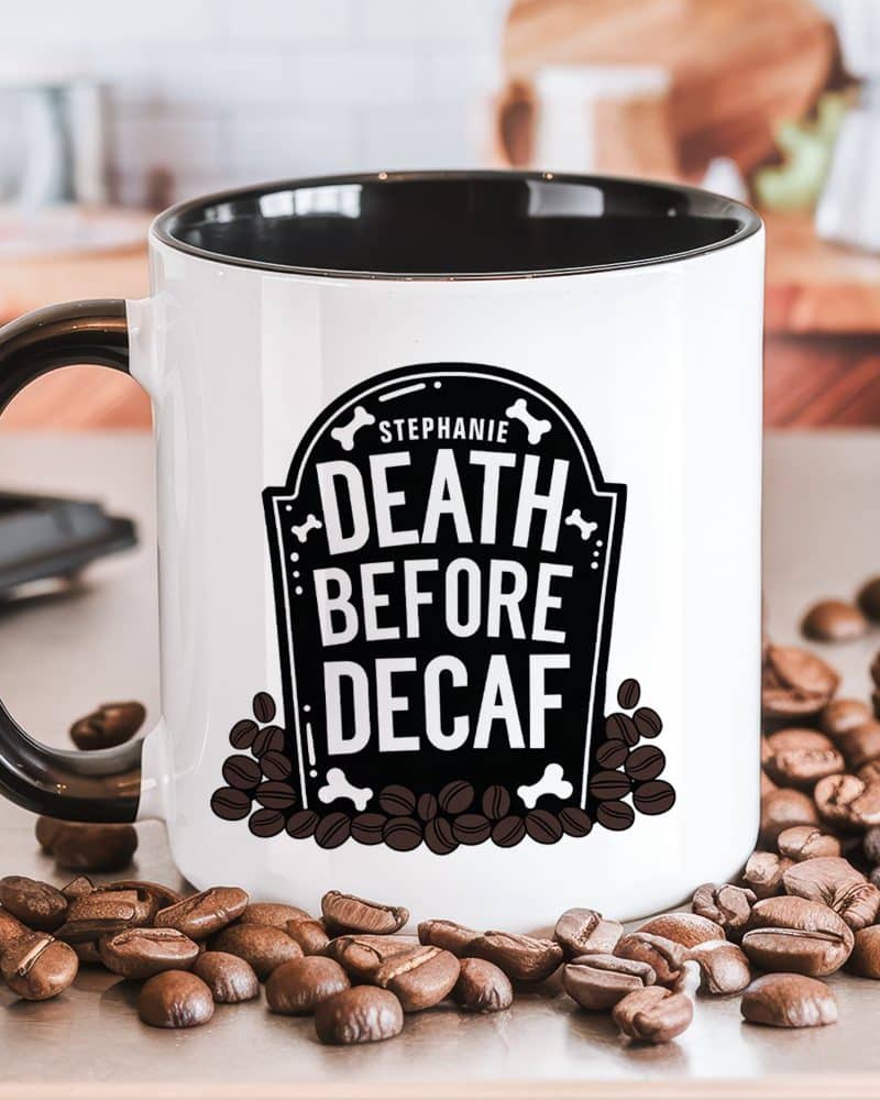 A white coffee mug with a black interior and handle, displaying the personalized text Stephanie, Death Before Decaf, within a tombstone graphic surrounded by coffee beans.