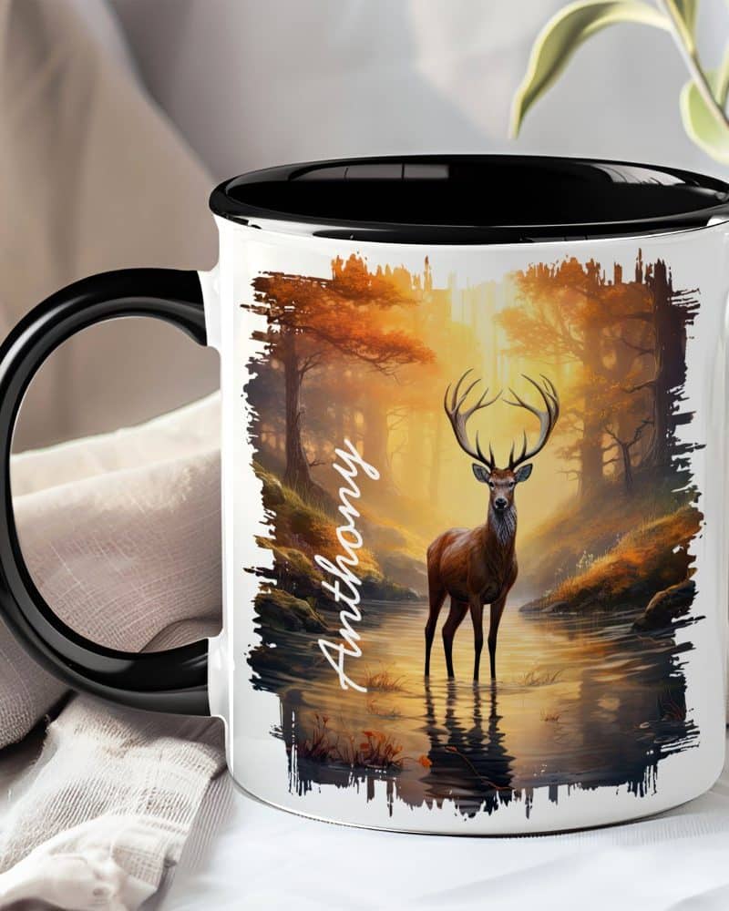 A coffee mug with a black handle and rim features a stag standing in a forest stream at sunset. The name Anthony is printed on the mug.
