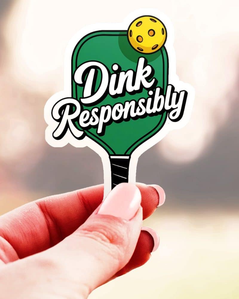 A hand holds a sticker of a pickleball paddle with the text Dink Responsibly.