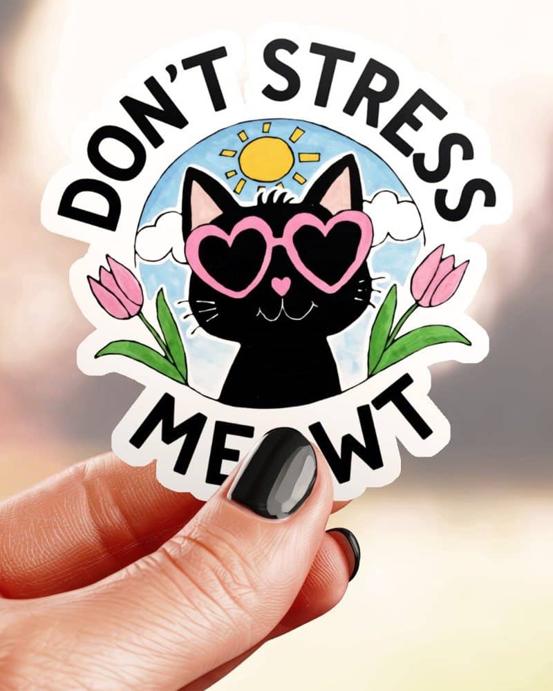 A hand holds a sticker featuring a black cat wearing heart-shaped sunglasses, surrounded by flowers and a sun, with the text Dont Stress Meowt.