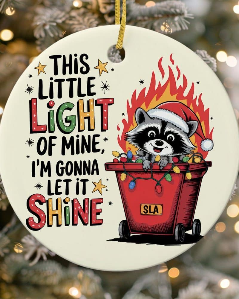 Christmas ornament featuring a raccoon wearing a Santa hat in a red container with Christmas lights, surrounded by flames. Text reads "This Little Light of Mine, I'm Gonna Let it Shine".