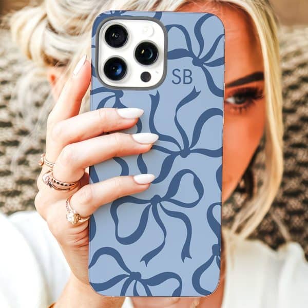 A Woman'S Hand Holds Up A Light Blue Iphone Case Featuring A Pattern Of Dark Blue Bows. The Initials Sb Are Visible On The Case.