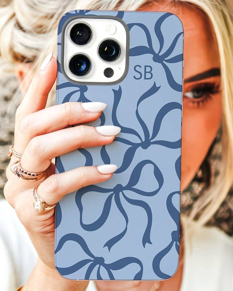 A woman's hand holds up a light blue iPhone case featuring a pattern of dark blue bows. The initials SB are visible on the case.