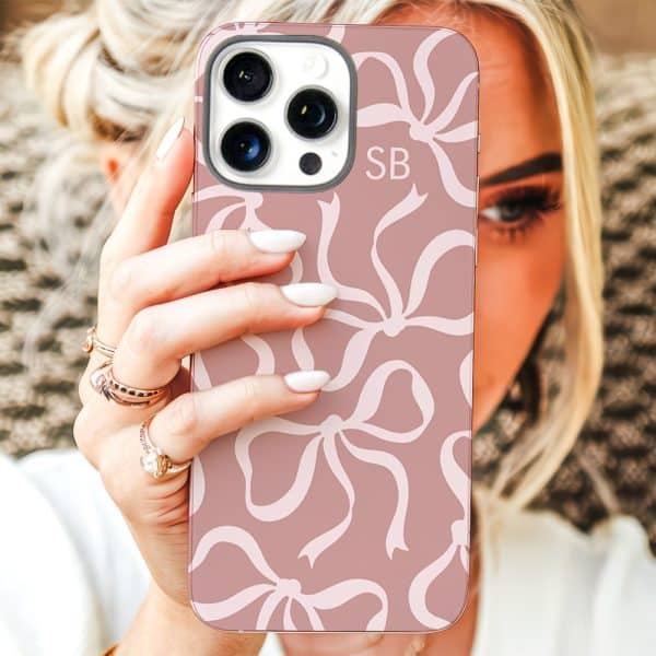 A Woman'S Hand Holds Up A Blush Pink Iphone Case Featuring A Repeating Pattern Of White Bows. The Initials Sb Are Visible On The Case.