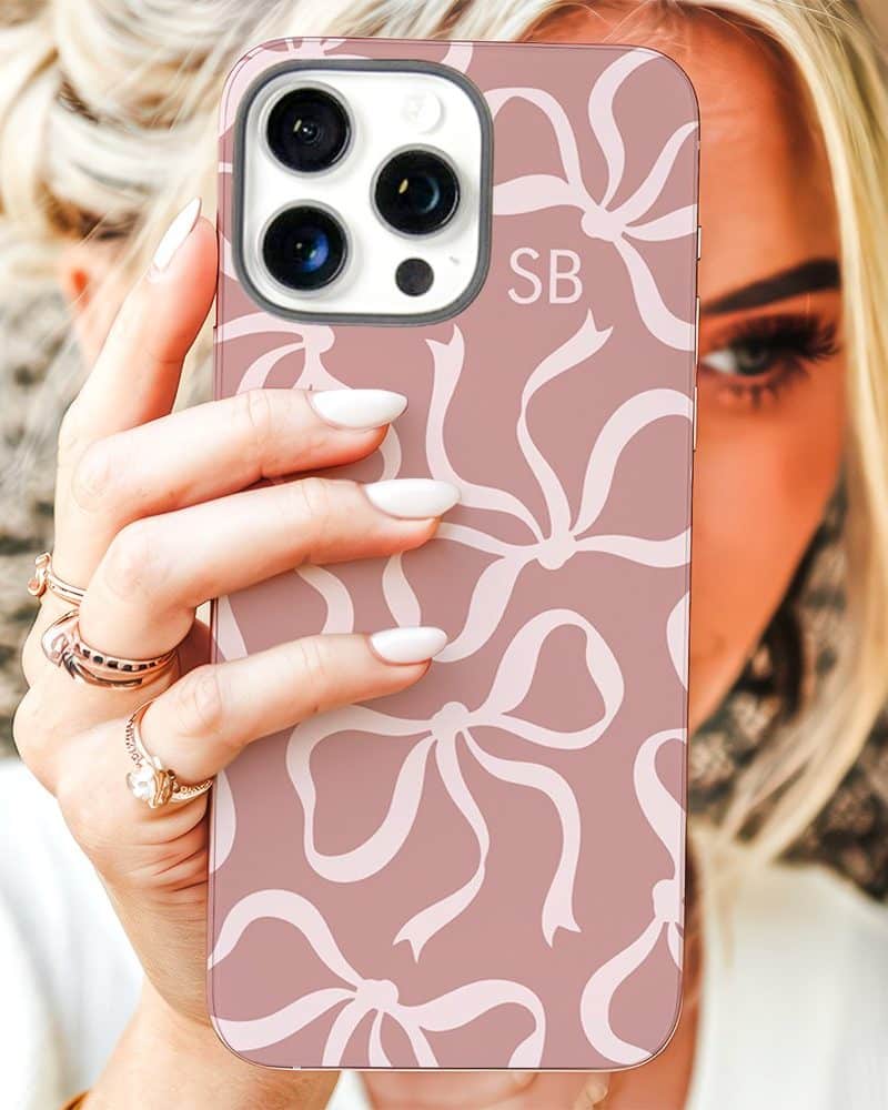A woman's hand holds up a blush pink iPhone case featuring a repeating pattern of white bows. The initials SB are visible on the case.