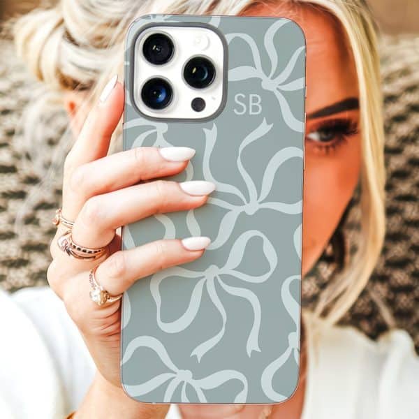 A Woman'S Hand Holds Up A Light Teal Phone Case Patterned With White Bows. The Case Features The Initials Sb.