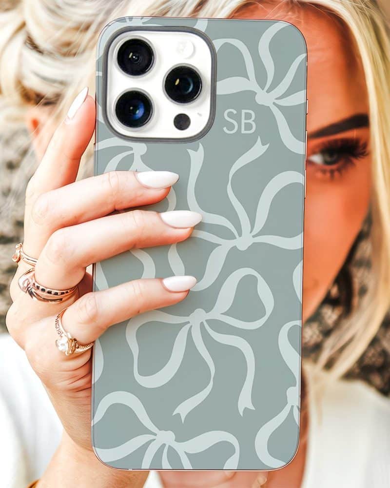 A woman's hand holds up a light teal phone case patterned with white bows. The case features the initials SB.