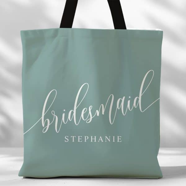 A Teal Tote Bag With The Word Bridesmaid In A Script Font And The Name Stephanie Underneath.