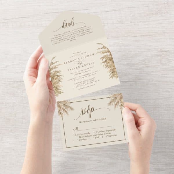 Two Hands Hold A Folded Wedding Invitation With Pampas Grass Design. The Invitation Includes The Couple'S Names And Wedding Details, Plus An Rsvp Card.