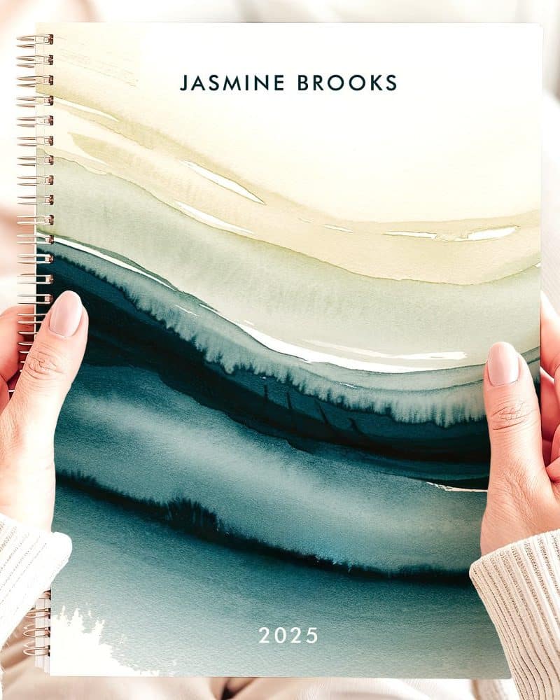 Two hands hold a 2025 spiral-bound planner featuring an abstract watercolor design in shades of teal and beige.