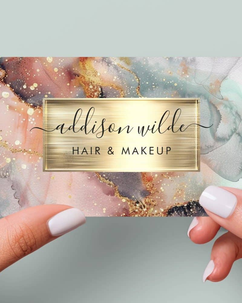 A business card for Addison Wilde Hair & Makeup, featuring an abstract watercolor design in blush pink, gold, and gray.