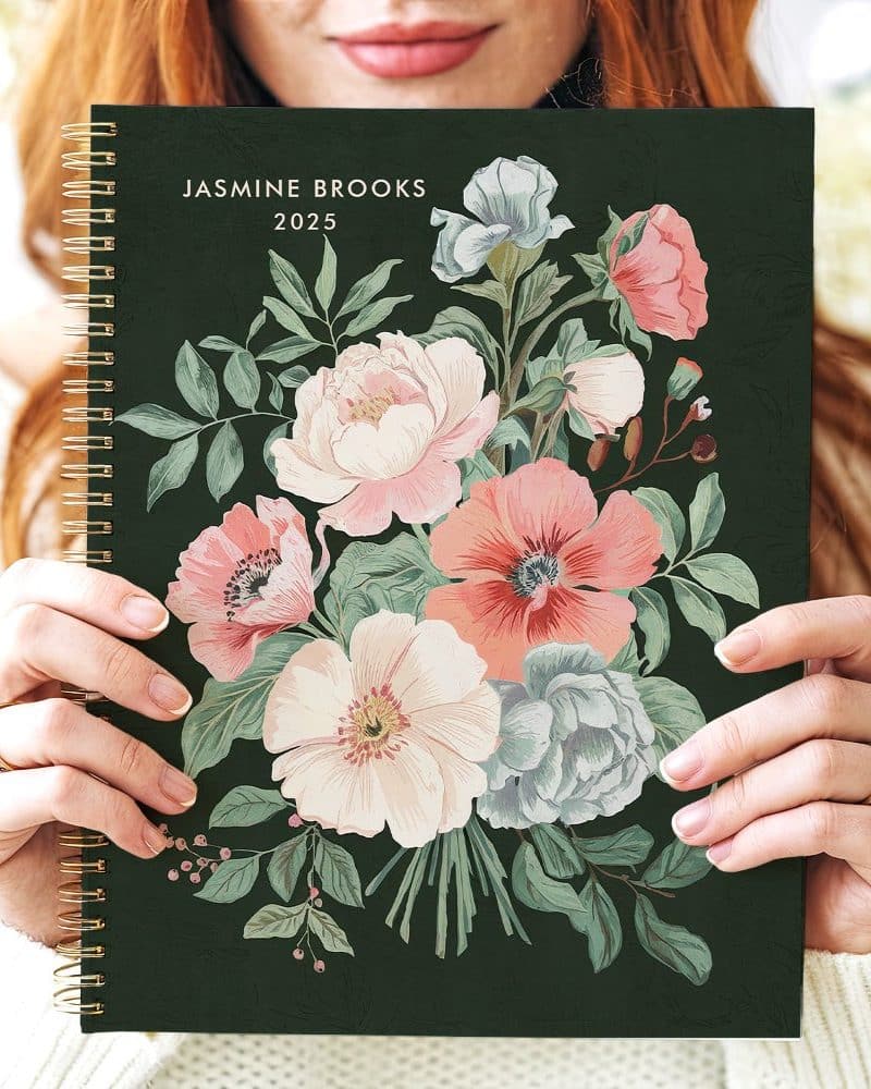 A woman holds a 2025 spiral-bound planner featuring a floral design on a dark green cover.