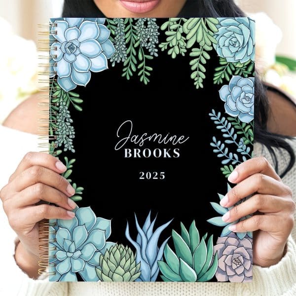 Person Holding A 2025 Planner With A Black Cover And A Border Of Illustrated Succulents.