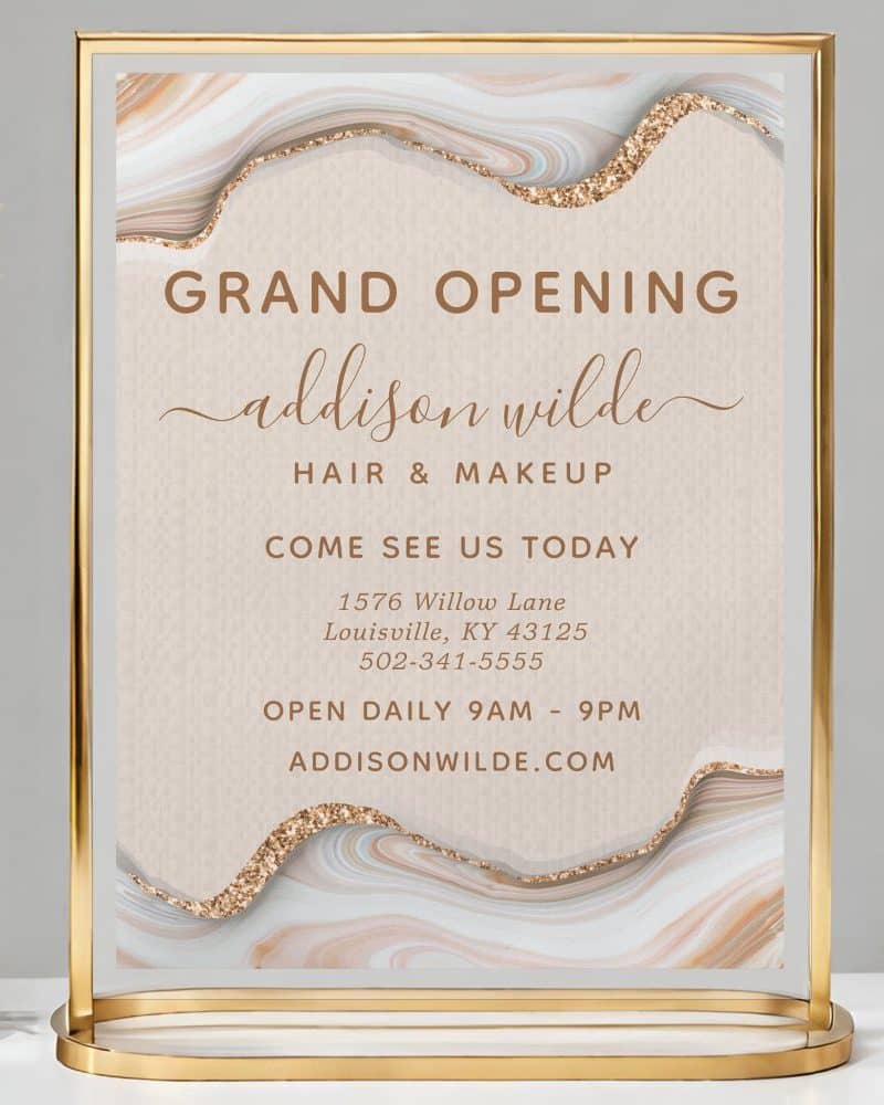 Gold stand-up sign announcing the grand opening of Addison Wilde Hair & Makeup, including address, phone number, and website.