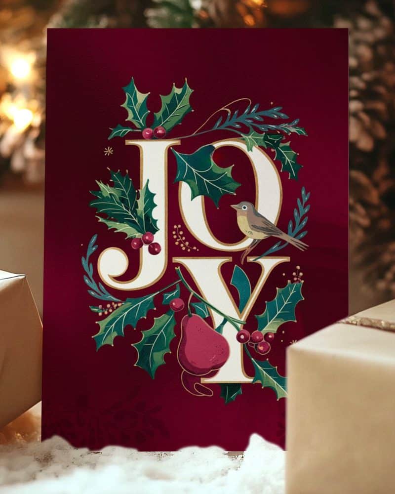 A burgundy Christmas card with the word JOY in elegant lettering surrounded by holly, berries, and a small bird.