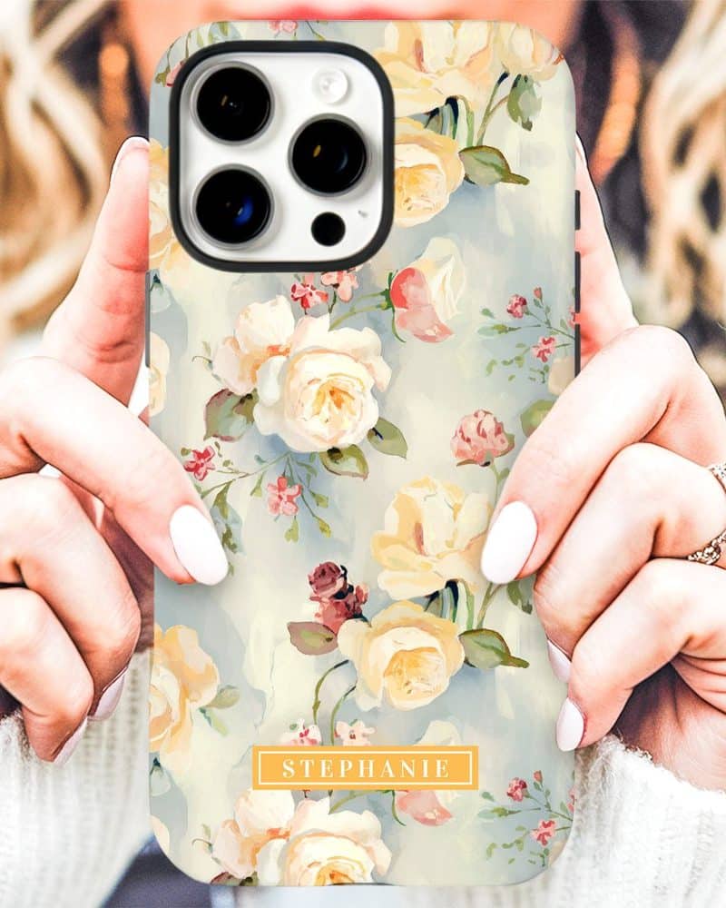 A woman's hands hold a phone case featuring a watercolor floral pattern of pale yellow roses and smaller flowers on a light blue background. The name Stephanie is printed on the case.