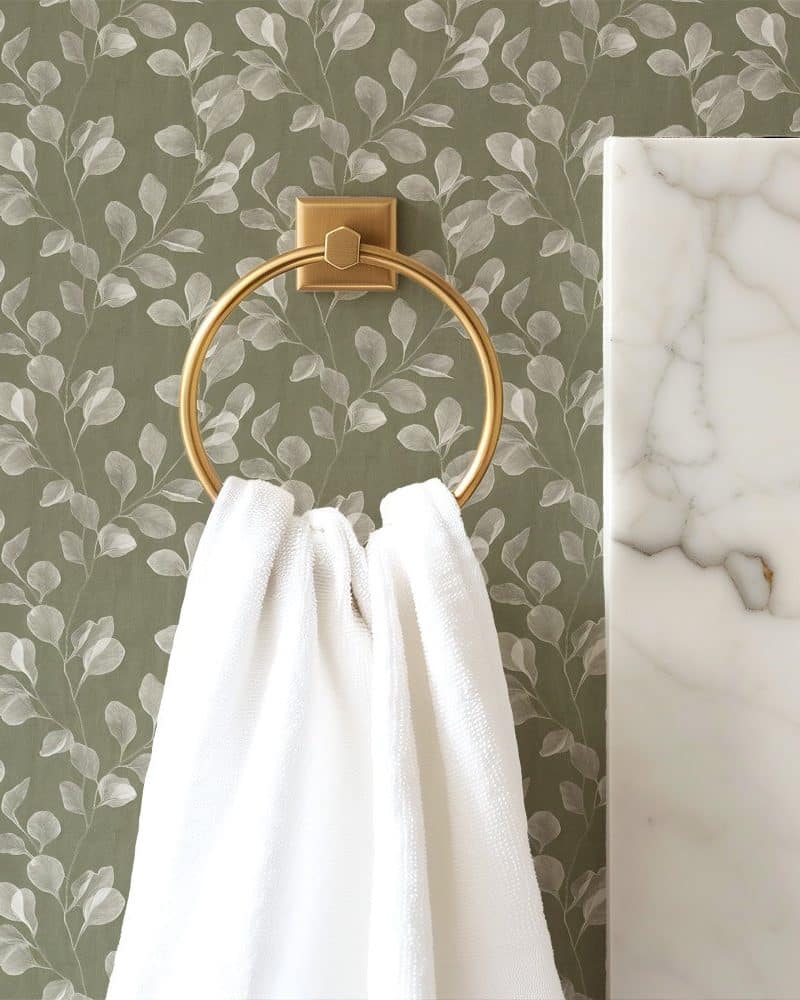 White towel hanging on a gold towel ring against a green leaf patterned wall.