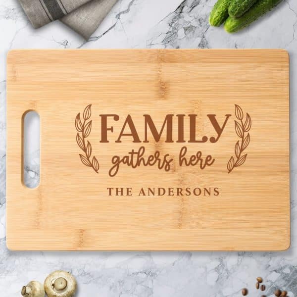 Light Brown Bamboo Cutting Board With The Words Family Gathers Here And The Andersons Engraved On It.