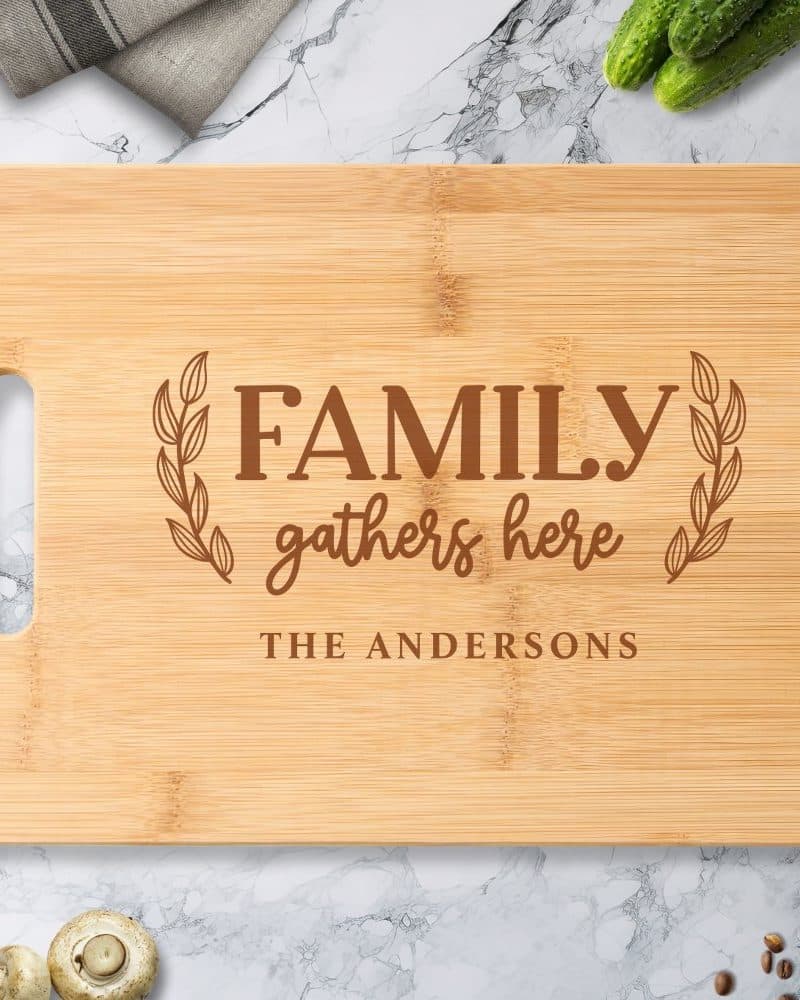 Light brown bamboo cutting board with the words Family gathers here and The Andersons engraved on it.