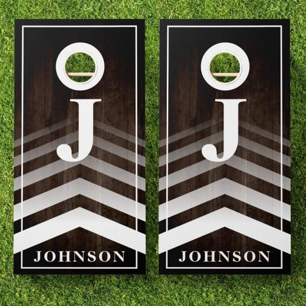 Pair Of Personalized Cornhole Boards With A Dark Wood Grain Background, Chevron Pattern, And Large White J Initial, Along With The Name Johnson.