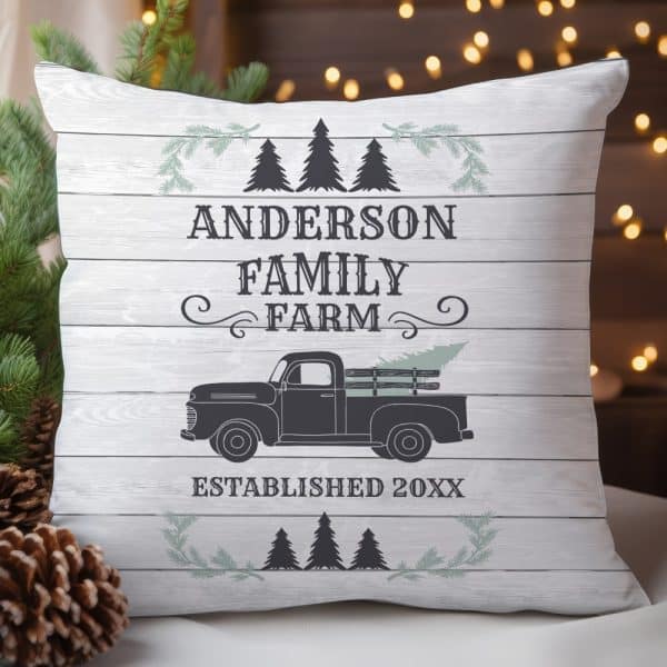 White Pillow With A Wood Grain Background Featuring The Anderson Family Farm Logo, Established Date, And A Vintage Truck Carrying A Christmas Tree.