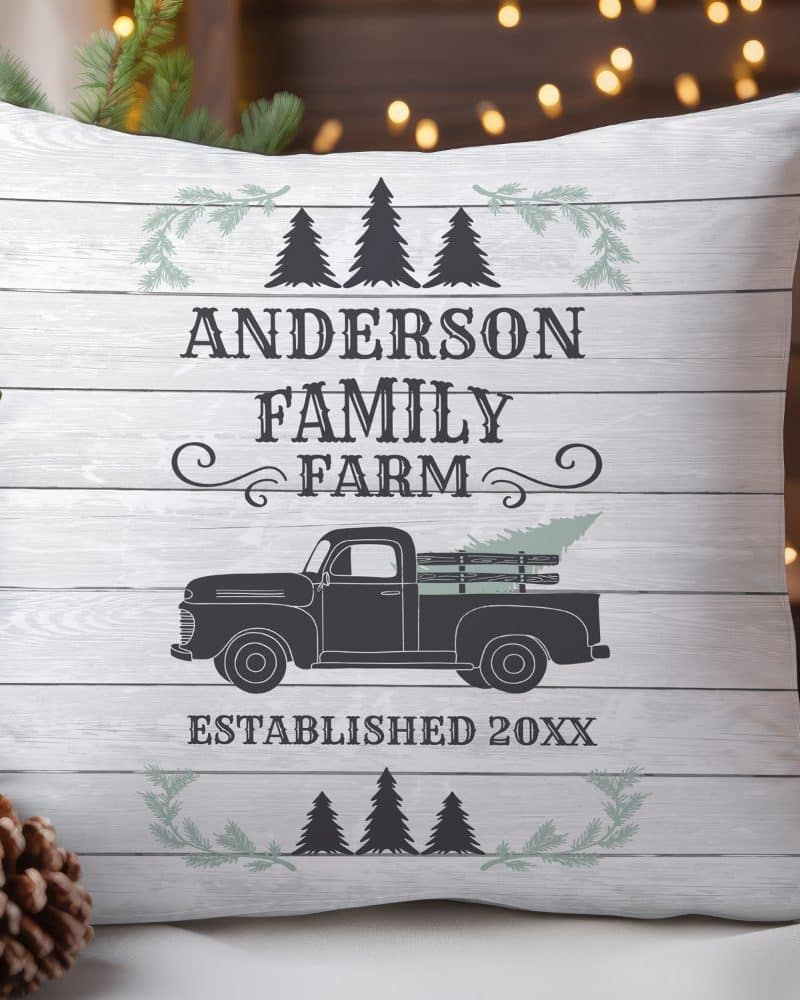White pillow with a wood grain background featuring the Anderson Family Farm logo, established date, and a vintage truck carrying a Christmas tree.