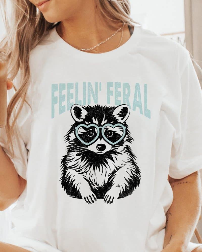 White t-shirt featuring a raccoon wearing heart-shaped sunglasses. Text above reads Feelin' Feral.