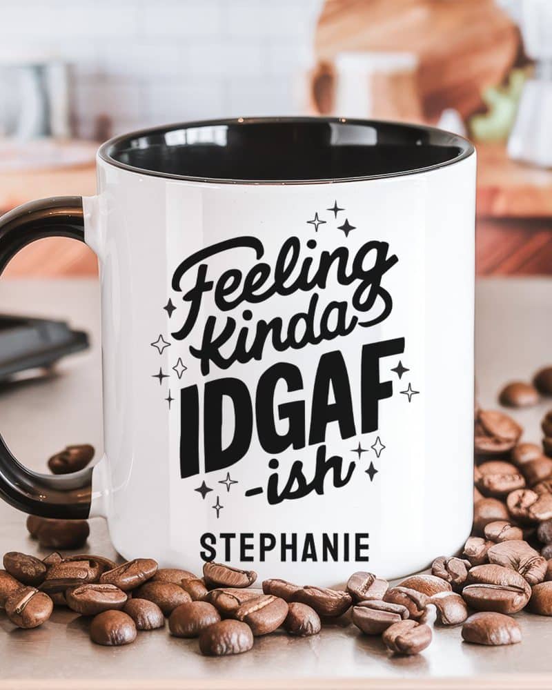 White coffee mug with black interior and handle, featuring the text Feeling Kinda IDGAF -ish and the name Stephanie. Coffee beans are scattered around the mug.