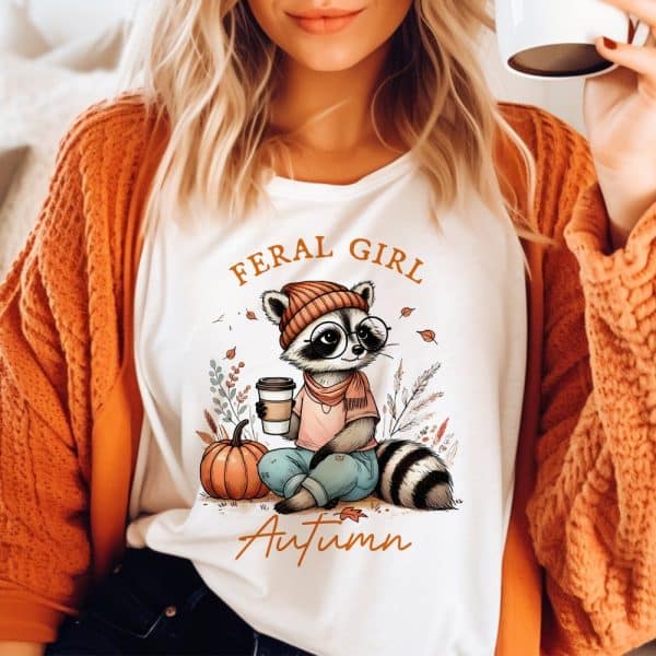 White Shirt Featuring A Cartoon Raccoon Wearing A Hat And Scarf, Holding A Coffee Cup, Sitting Next To A Pumpkin. Text Reads Feral Girl Autumn.