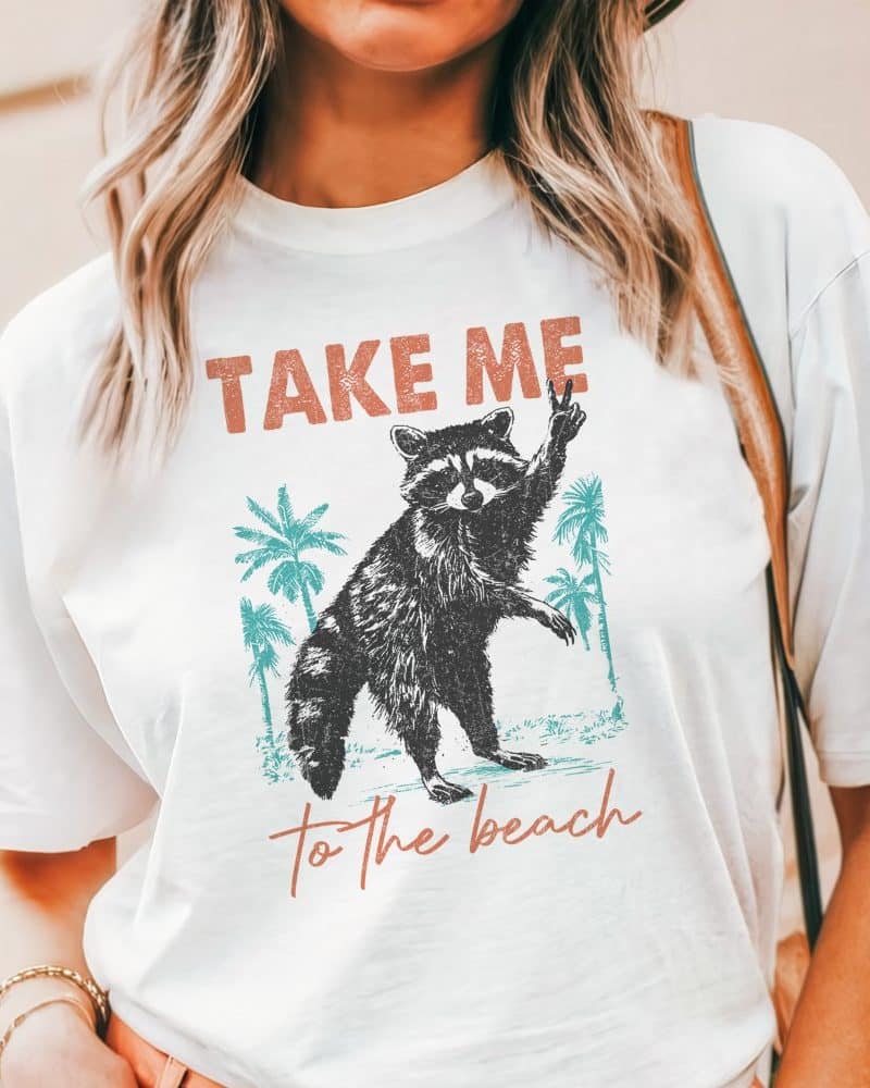 White t-shirt featuring a graphic design of a raccoon giving a peace sign, palm trees, and the text Take Me To The Beach.