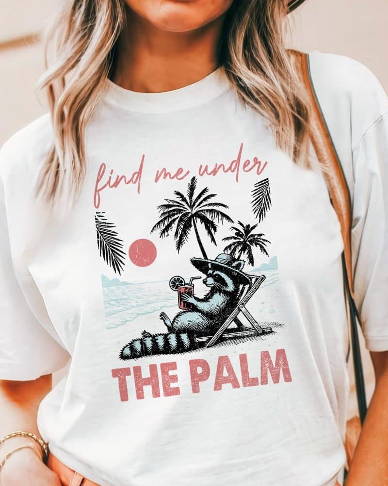 White t-shirt featuring a raccoon lounging in a beach chair under palm trees, holding a drink. Text reads Find Me Under The Palm.