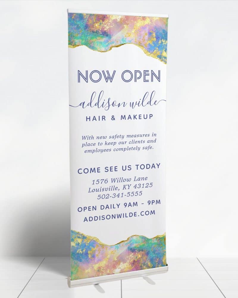 A retractable banner announcing the grand opening of Addison Wilde Hair & Makeup. The banner features a pastel abstract design and includes the business's address, phone number, and website.