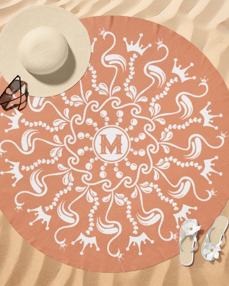 Peach-colored round beach towel with a white mandala design featuring crowns and the letter M in the center.