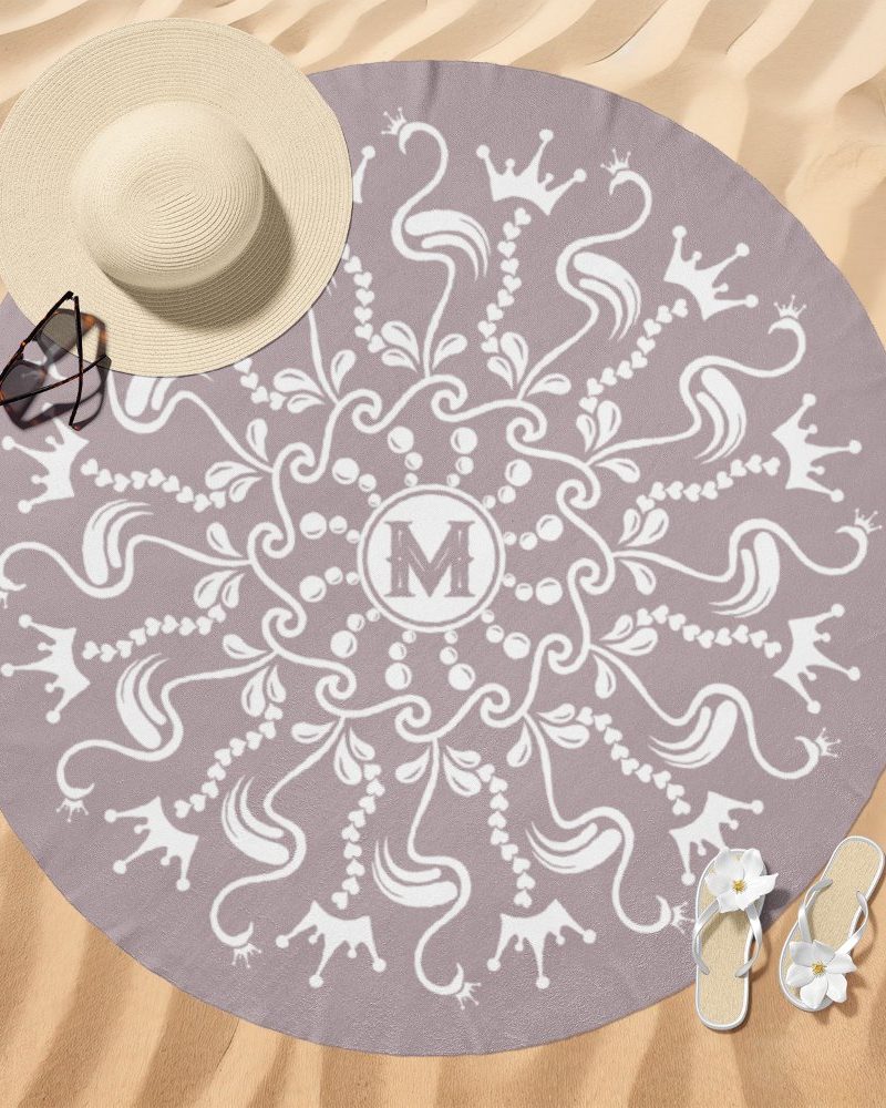 A mauve round beach towel with a white mandala design featuring crowns and the letter M in the center.
