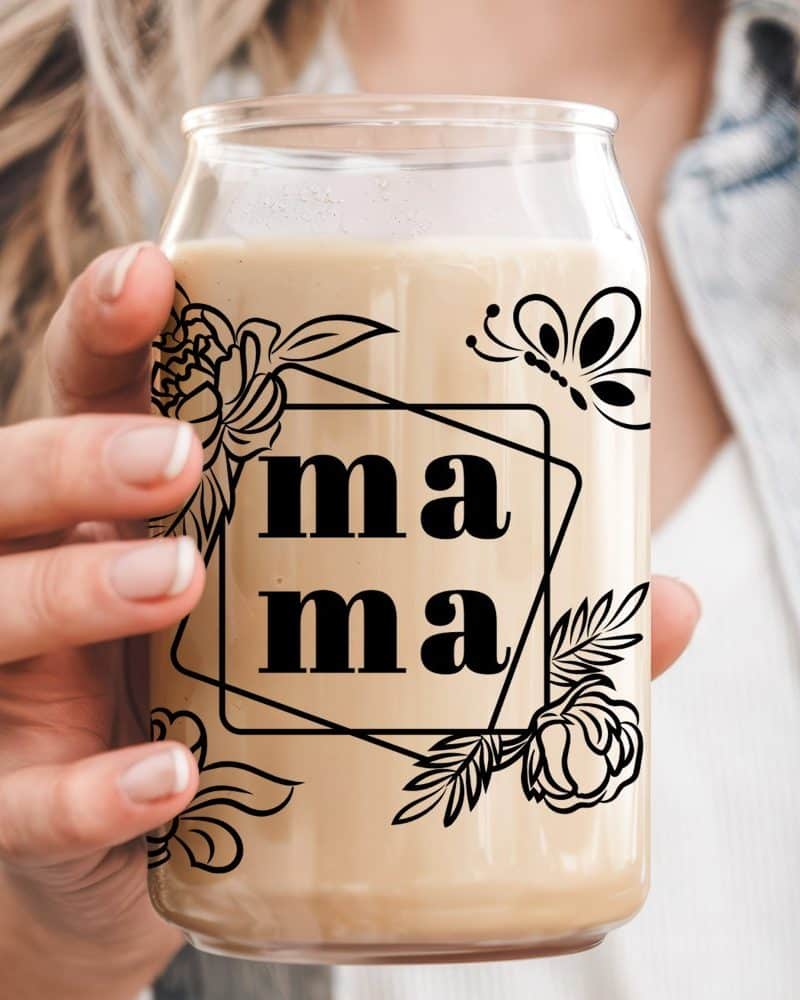 A hand holds a glass with a beige beverage. The glass features a design of the word mama, flowers, and a butterfly.