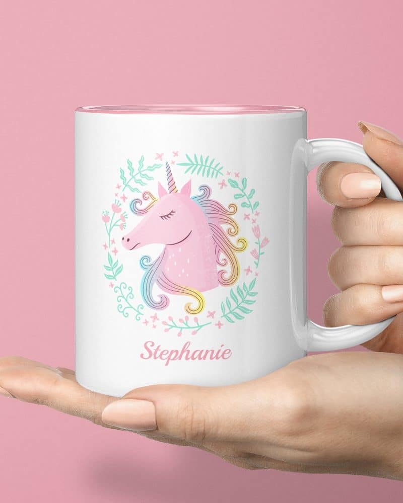 A hand holds a white mug featuring a pastel pink unicorn surrounded by flowers and the name Stephanie.