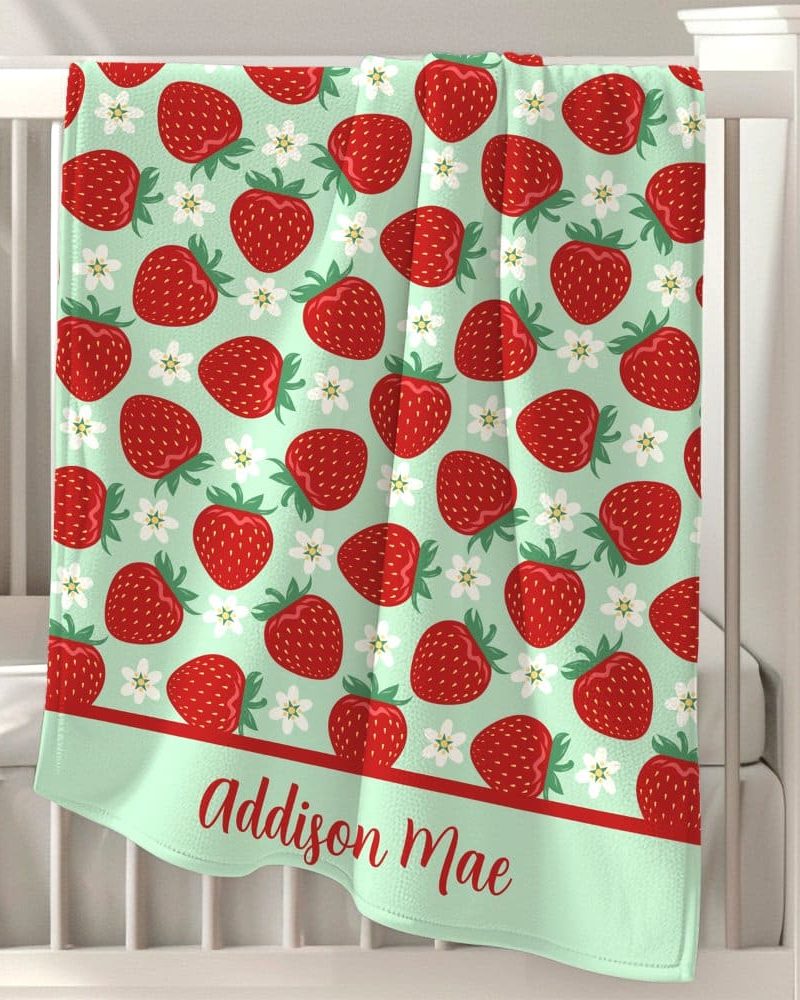 A light green baby blanket with a repeating pattern of red strawberries and white flowers, personalized with the name Addison Mae.