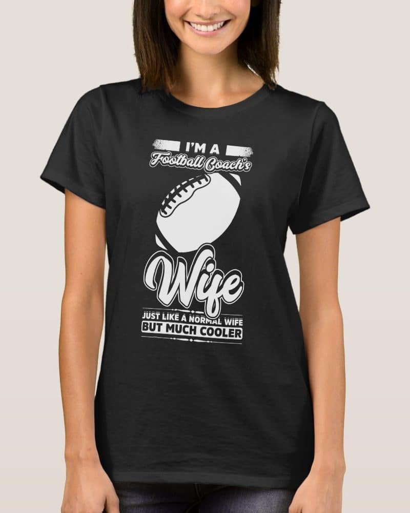 Woman wearing a black t-shirt that says Im a Football Coachs Wife, with a football graphic and the phrase Just like a normal wife but much cooler.